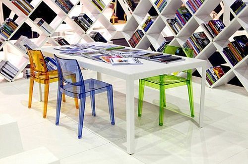 Estantes geométricas com livros coloridos e cadeiras de acrílico.